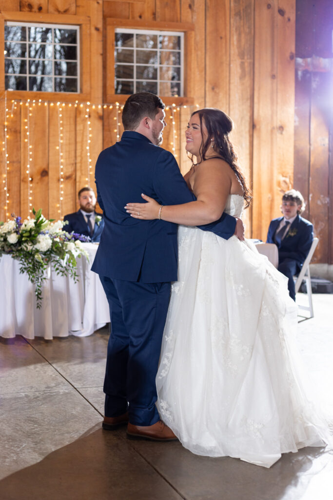 The bride and groom share their first dance inside the reception space during their chattanooga barn wedding, holding each other closely on the concrete floor as warm string lights glow against the wooden walls and guests watch from nearby tables.
