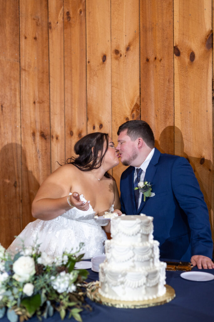 The couple shares a playful kiss after feeding each other cake during their chattanooga barn wedding, the bride holding a fork as the groom leans in, with the textured white cake and wooden barn wall filling the frame.