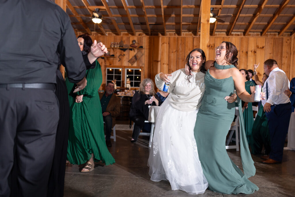 The bride dances with a bridesmaid during the reception of their chattanooga barn wedding, laughing with arms around each other as guests sit and watch from white chairs in the background.