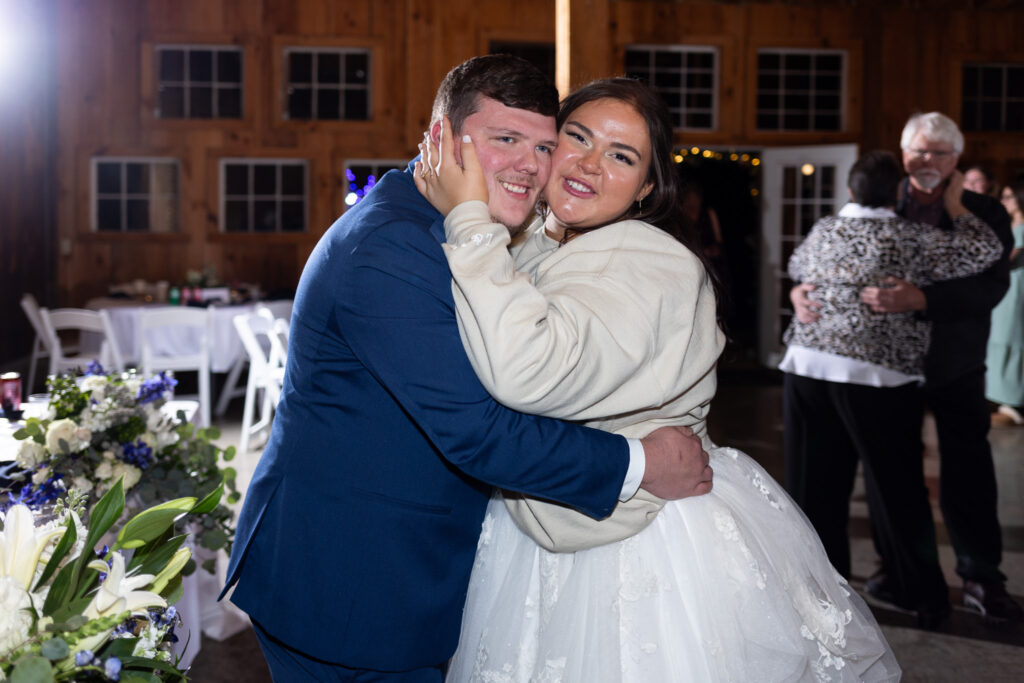 A close-up reception portrait shows the bride and groom embracing during their chattanooga barn wedding, the bride wearing a cozy sweatshirt over her wedding dress as they smile directly at the camera on the dance floor.