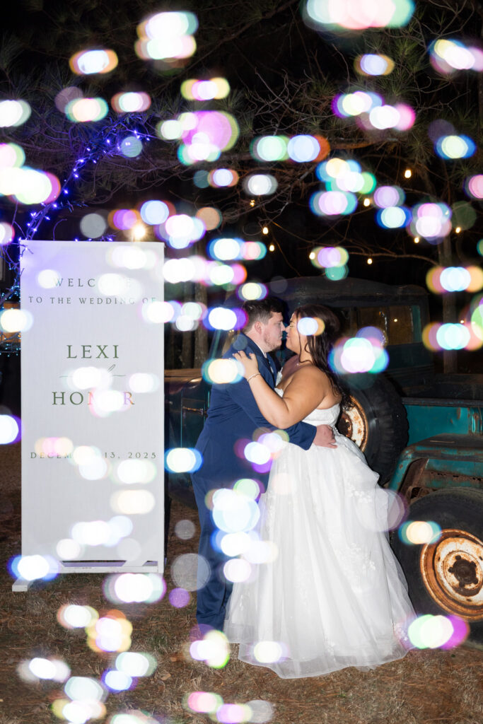 The bride and groom embrace beneath colorful bokeh lights during their chattanooga barn wedding, standing beside a vintage truck as a welcome sign and glowing string lights create a dreamy nighttime backdrop.