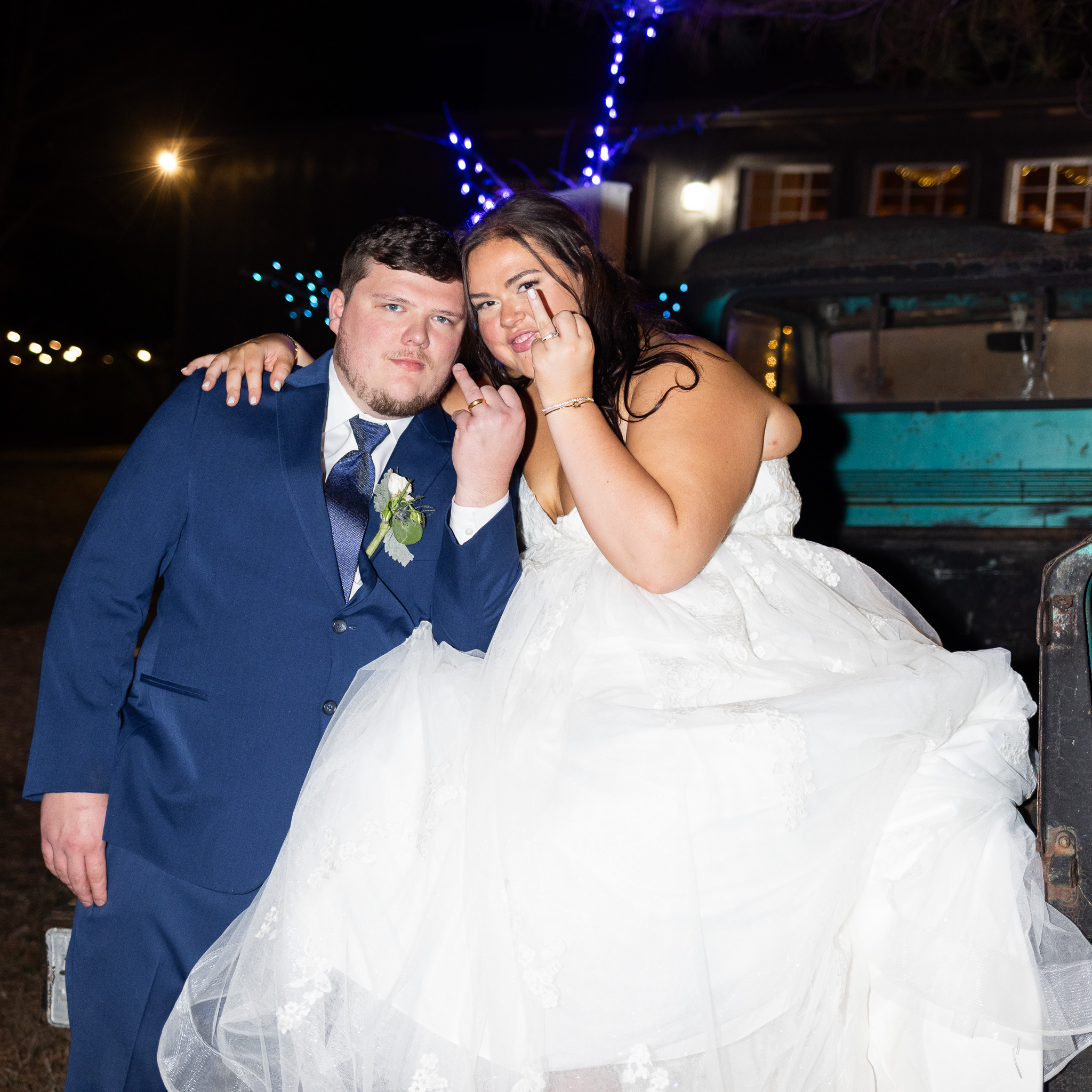 The bride and groom sit closely together for a nighttime portrait during their chattanooga barn wedding, holding up their wedding rings toward the camera while leaning against a vintage truck decorated with string lights.