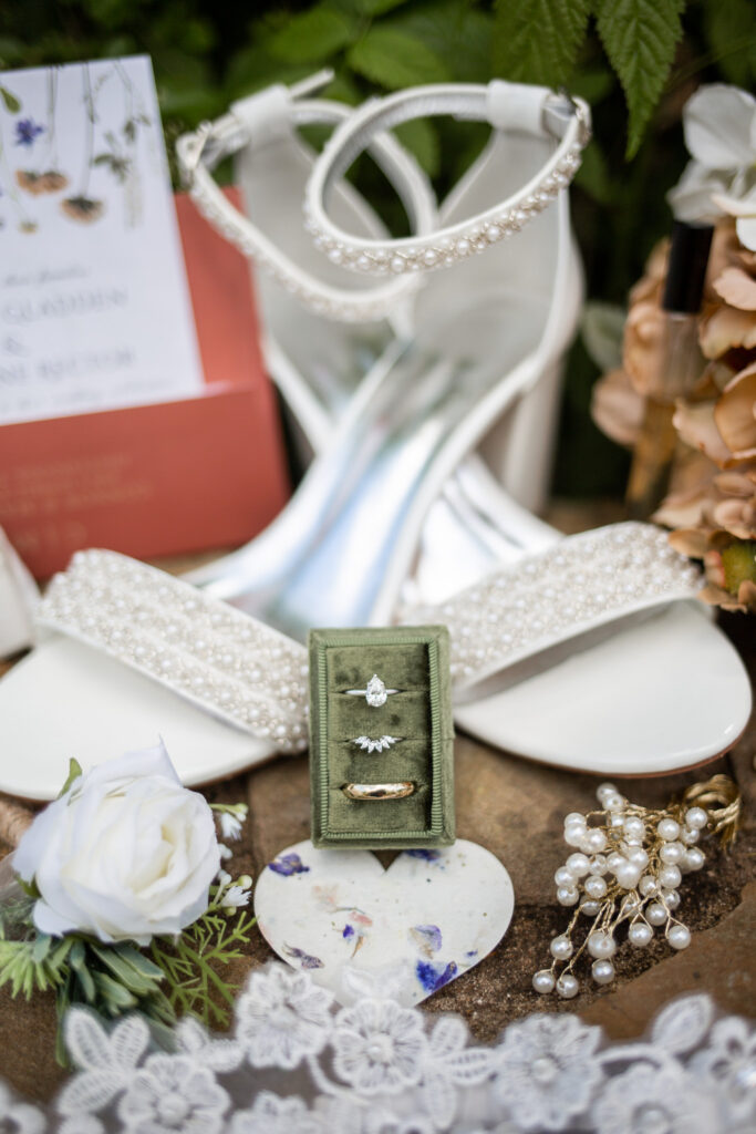 Wedding detail photos featuring a green velvet ring box holding three rings arranged vertically, placed on a textured stone surface and surrounded by white floral accents, pearl jewelry, lace trim, and beaded white bridal heels positioned softly in the background with greenery framing the scene.