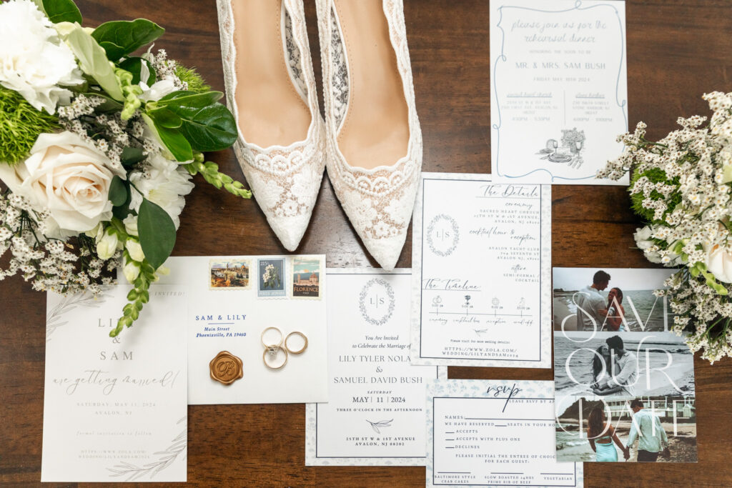 Flat lay wedding detail photos showcasing lace pointed-toe bridal shoes centered at the top of a wooden surface, with a full invitation suite arranged below, including envelopes, stamps, RSVP cards, and rings, balanced by loose greenery and white florals placed symmetrically on either side.