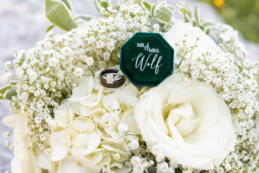 Wedding detail photos highlighting a green velvet hexagon ring box embroidered with “Mr & Mrs Wolf,” resting gently inside a lush white bouquet filled with hydrangeas and baby’s breath, with wedding rings nestled beside the box among soft petals and natural textures.
