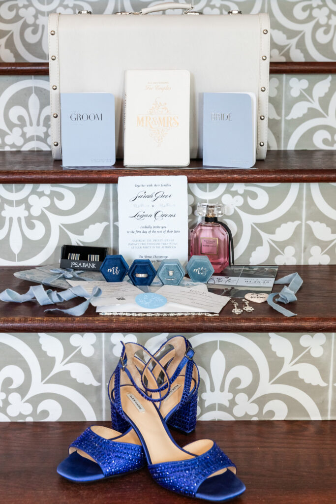 Wedding detail photos arranged on a tiled staircase shelf featuring a white bridal clutch at the top, vow books labeled groom and bride, an invitation suite, blue velvet ring boxes, jewelry, ribbon, perfume, and deep blue beaded heels placed on the lower step for a layered, editorial look.