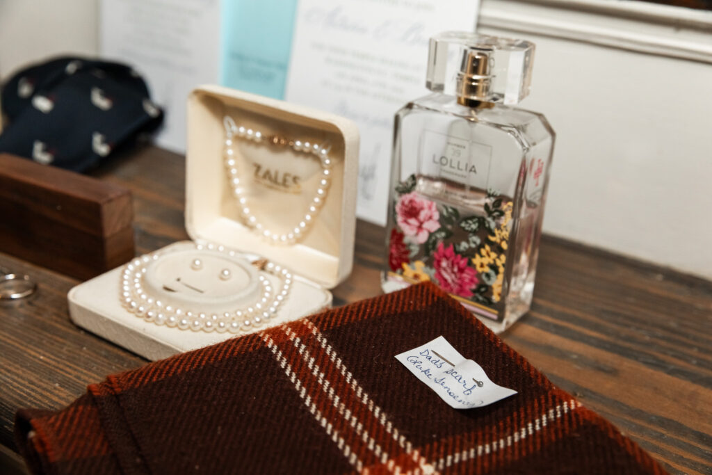 Wedding detail photos capturing heirloom jewelry displayed inside a wooden box lined with pearls, alongside a floral-labeled perfume bottle and a folded plaid fabric with a handwritten note pinned to it, arranged on a dark wooden surface that emphasizes warmth and sentimental value.