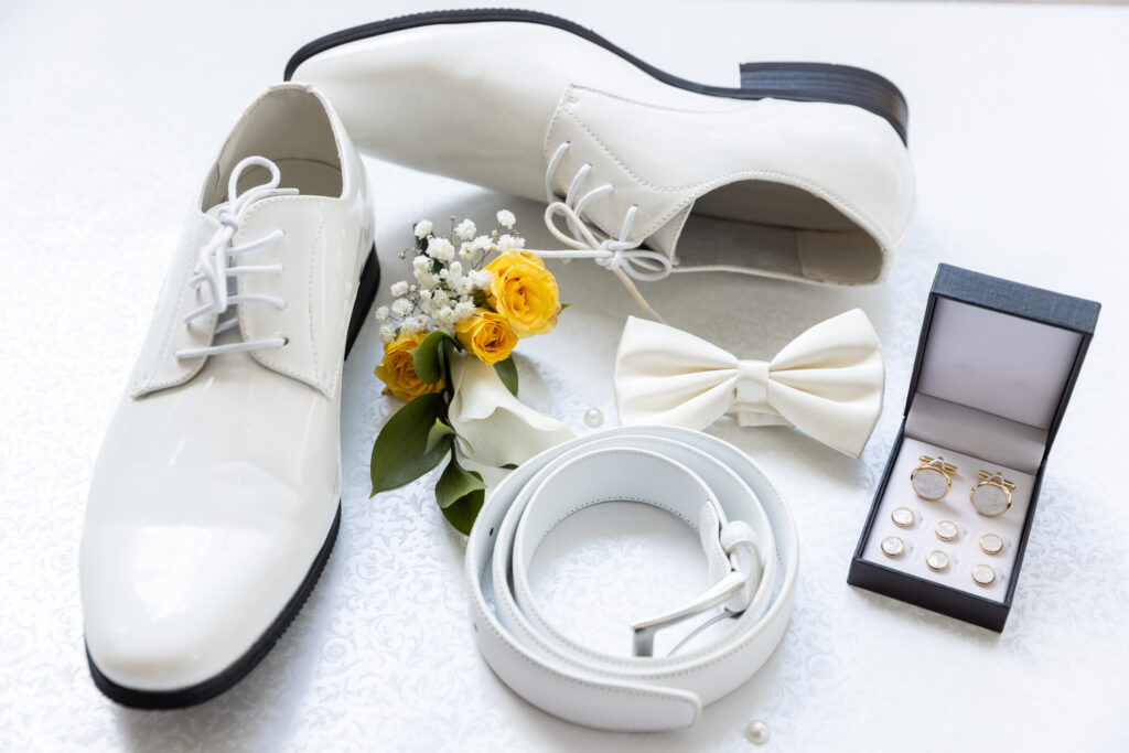 Wedding detail photos featuring white groom’s dress shoes styled with a matching belt, cufflinks in a black box, a cream bow tie, and a small yellow and white floral boutonniere, all arranged neatly on a bright textured surface with soft, even lighting.