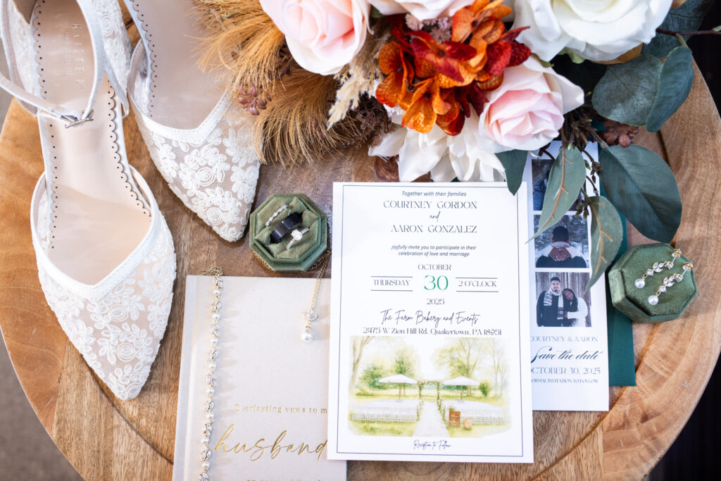 Wedding flat lay on a round wooden table featuring lace bridal heels, a bouquet with blush and rust-toned florals, an invitation suite with save-the-date cards, vow book, ring boxes, earrings, and greenery arranged to highlight texture, color, and personal details.