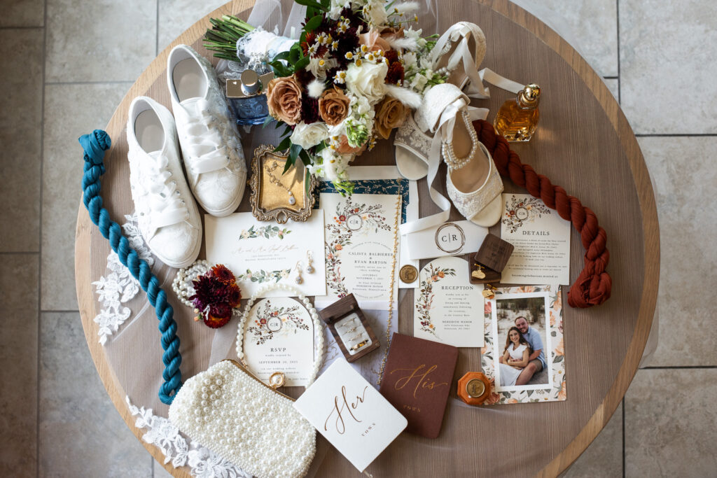 Overhead wedding flat lay styled on a round wooden table, showcasing a full invitation suite, vow books labeled his and hers, rings, jewelry, braided fabric elements, lace, perfume bottles, bridal shoes, and a bouquet, arranged intentionally to reflect color, texture, and personal touches.