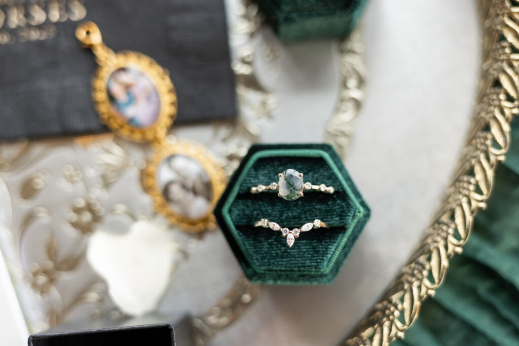 Detailed flat lay of a green velvet ring box holding a non-traditional engagement ring and wedding band, styled alongside heirloom photo lockets, gold jewelry, textured fabric, and soft neutral tones arranged on a decorative surface.