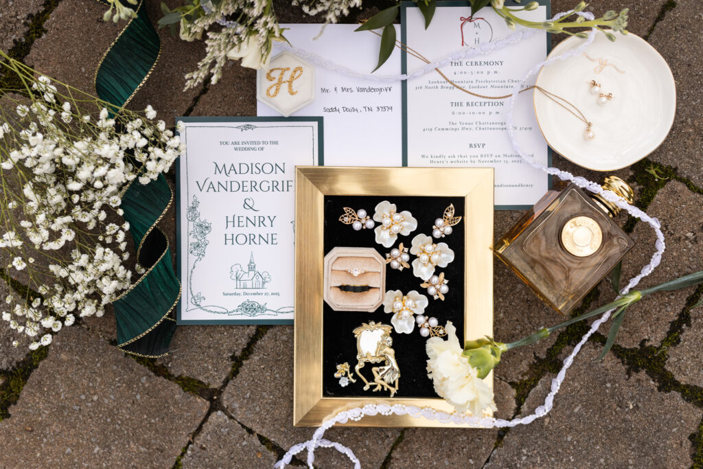 Overhead wedding flat lay arranged on brick, showcasing a full invitation suite with ceremony and reception cards, a gold-framed tray holding wedding rings and floral jewelry, pearl accessories, loose greenery, ribbon, and a glass perfume bottle placed among soft white florals.