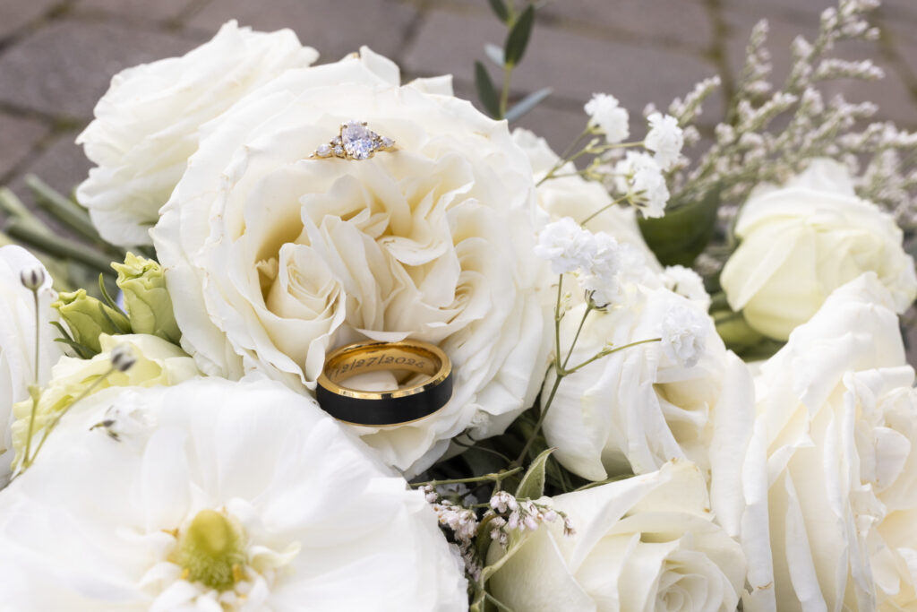 A close-up detail of the wedding rings nestled into the center of a full white rose bouquet, with the engagement ring resting atop layered petals and the wedding band tucked below, surrounded by additional roses and airy sprigs of baby’s breath that fill the frame with soft texture.