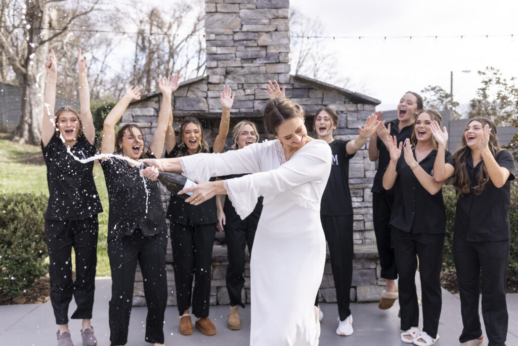 A celebratory champagne pop with the bride standing front and center in her getting-ready robe while her bridesmaids cheer behind her with raised arms, documenting the excitement and energy 