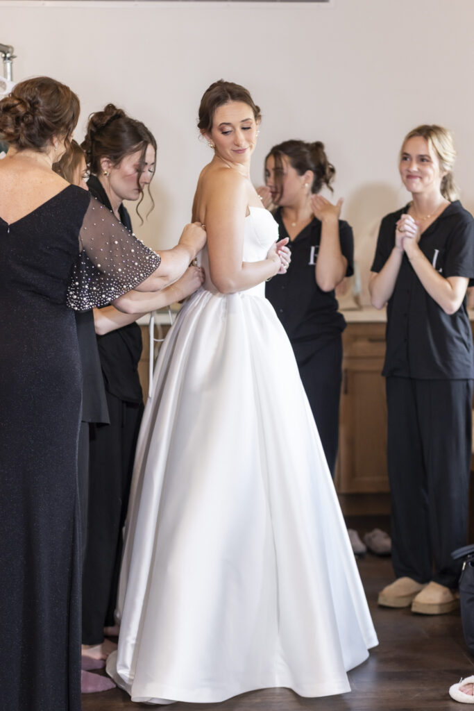 The bride stands in profile while her dress is carefully zipped by a family member, surrounded by bridesmaids in dark getting-ready outfits, their expressions warm and supportive during preparations for this Chattanooga Church Wedding.