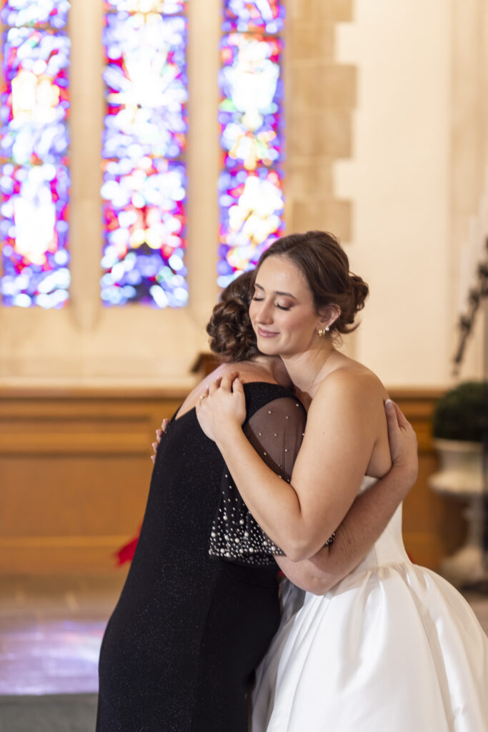 Inside the church, the bride embraces an older woman tightly, eyes closed and face peaceful, sunlight filtering through stained glass windows behind them in a quiet, emotional moment