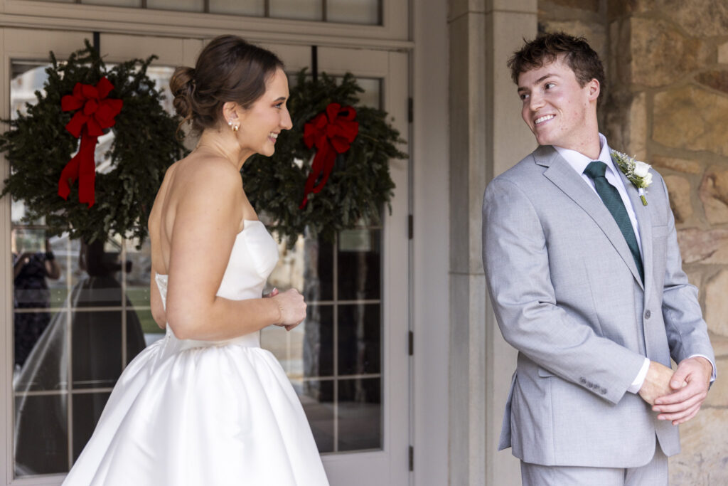A joyful first look moment outside the stone church, where the bride in a strapless satin gown approaches the groom from behind near glass doors decorated with winter wreaths, capturing the anticipation and emotion of a Chattanooga Church Wedding as he turns with a wide smile to see her for the first time.