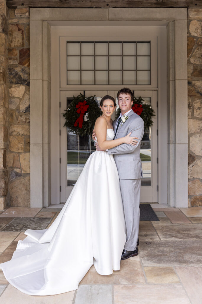 Outside the church entrance, the bride and groom stand together in front of glass doors decorated with wreaths, arms wrapped around one another, her long train trailing across the stone floor at the close of their Chattanooga Church Wedding.