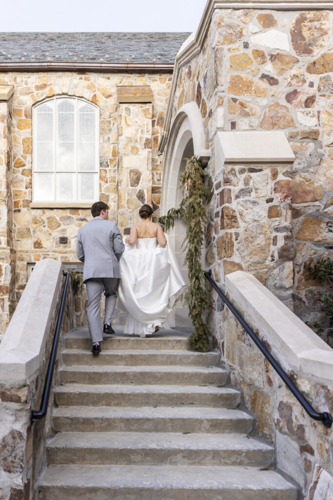 From behind, the bride and groom walk up stone steps toward a church entrance wrapped in greenery, her gown lifted slightly as they ascend together during their Chattanooga Church Wedding.