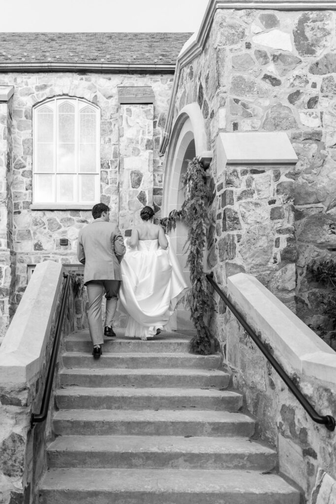 From behind, the bride and groom walk up stone steps toward a church entrance wrapped in greenery, her gown lifted slightly as they ascend together during their Chattanooga Church Wedding.