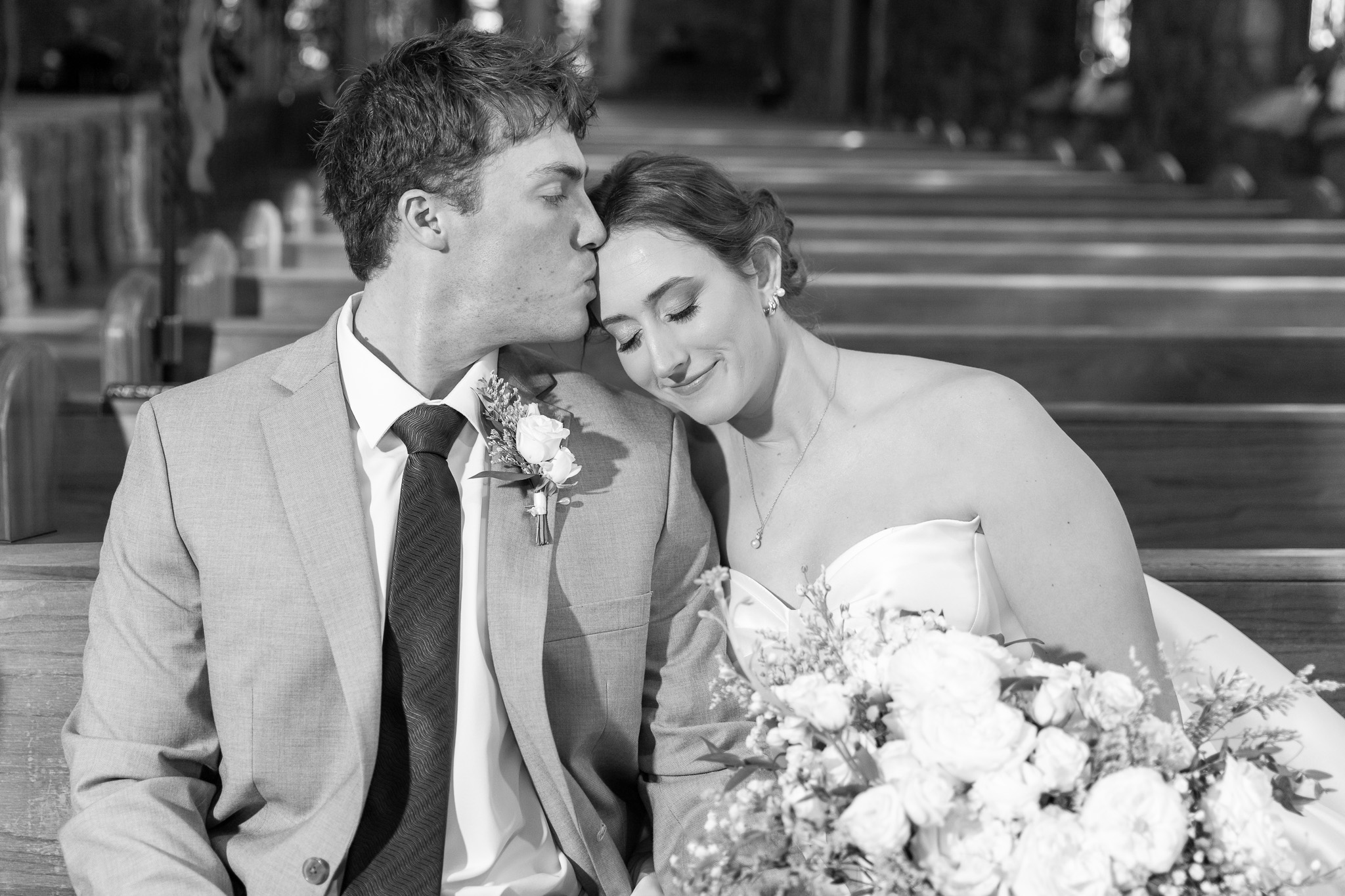 A black and white portrait of the groom leaning in to kiss the bride’s forehead as they sit together in a wooden church pew, her eyes closed and smiling softly, bouquet resting in her lap during their Chattanooga Church Wedding, with long rows of empty pews fading into the background.