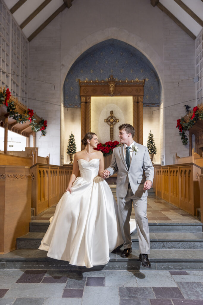 The couple walk hand in hand down the church steps inside the sanctuary, smiling at one another beneath arched stonework and a large cross, capturing a joyful moment from their Chattanooga Church Wedding ceremony.