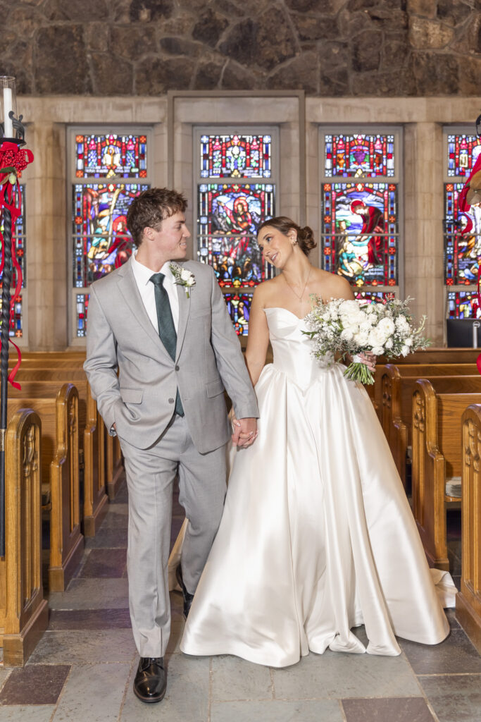 The bride and groom stroll between wooden pews, holding hands and gazing at each other, with detailed stained glass windows lining the aisle during their Chattanooga Church Wedding.
