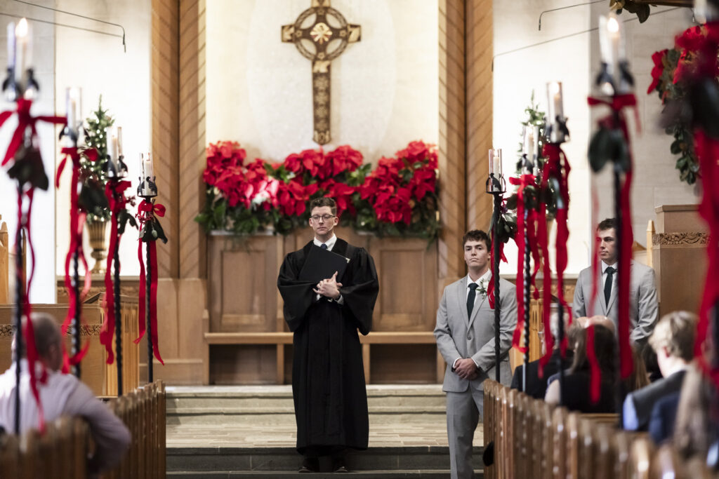 The officiant stands centered at the altar with the groom and groomsmen positioned to one side, all facing the congregation. Tall candles and greenery line the aisle, creating depth and symmetry leading toward the cross above. This scene sets the tone and structure of the Lookout Mountain Presbyterian Wedding, moments before the ceremony unfolds.