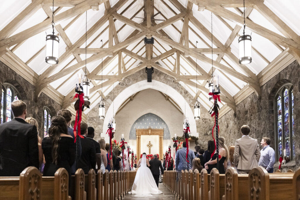 A wide view from the back of the church shows guests standing in the pews as the bride walks down the aisle toward the altar, framed by exposed beams, hanging lanterns, and stone walls during this Chattanooga Church Wedding.