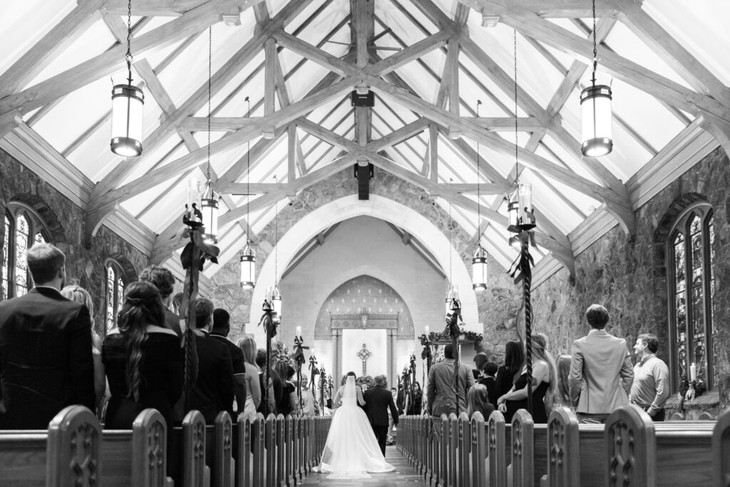 A wide view from the back of the church shows guests standing in the pews as the bride walks down the aisle toward the altar, framed by exposed beams, hanging lanterns, and stone walls during this Chattanooga Church Wedding.