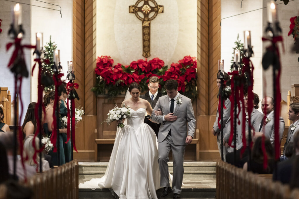 The couple walks back down the church aisle together after the ceremony, arms linked as they step forward in unison. Guests stand on either side of the pews, watching as the officiant follows behind them. The cross above the altar centers the frame, marking a meaningful milestone within their Lookout Mountain Presbyterian Wedding ceremony.