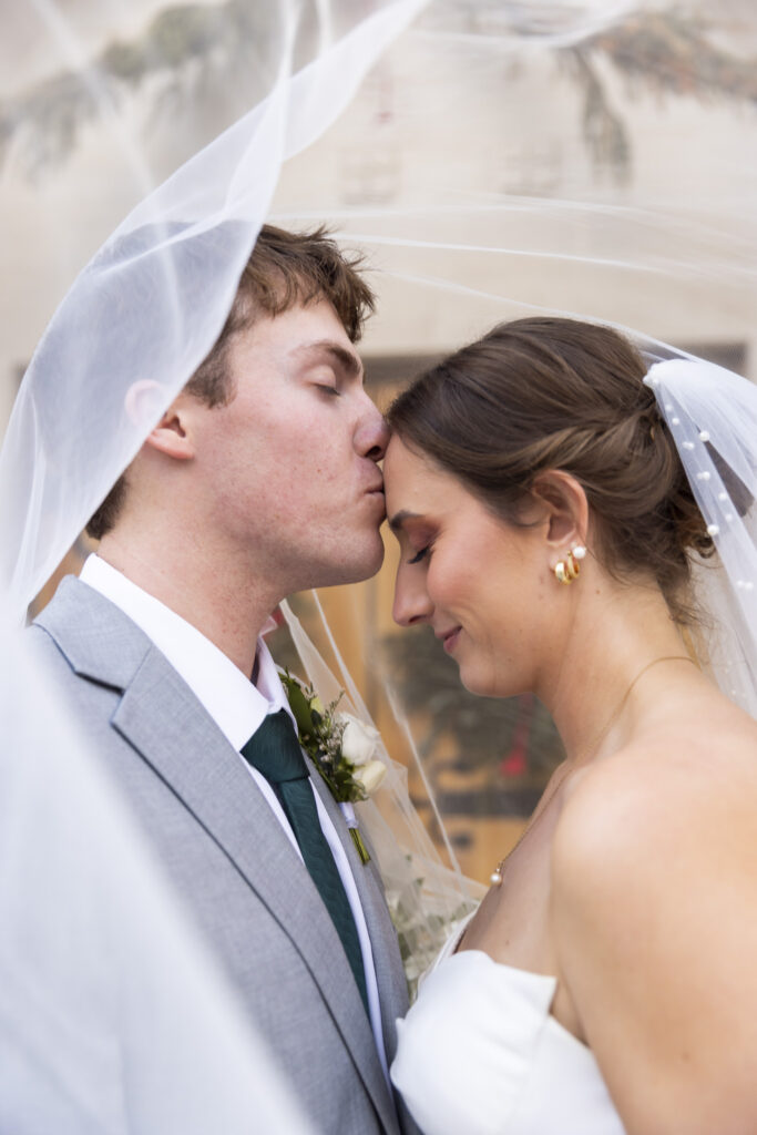 A close, emotional portrait beneath the bride’s veil as the groom kisses her forehead, both of their eyes closed. The veil drapes softly around their faces, creating a cocooned, private feeling despite the public setting. This quiet exchange adds emotional depth to their Lookout Mountain Presbyterian Wedding portraits.