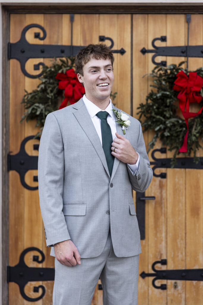 The groom stands alone in front of large wooden church doors decorated with greenery, smiling slightly as he adjusts his jacket near his boutonniere. His posture is relaxed but anticipatory, capturing a moment of calm before the ceremony. The setting places this portrait squarely within their Lookout Mountain Presbyterian Wedding day.