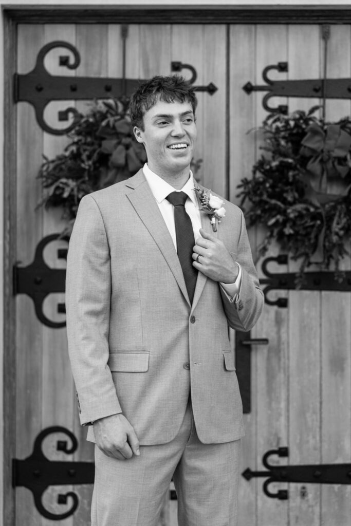 The groom stands alone in front of large wooden church doors decorated with greenery, smiling slightly as he adjusts his jacket near his boutonniere. His posture is relaxed but anticipatory, capturing a moment of calm before the ceremony. The setting places this portrait squarely within their Lookout Mountain Presbyterian Wedding day.