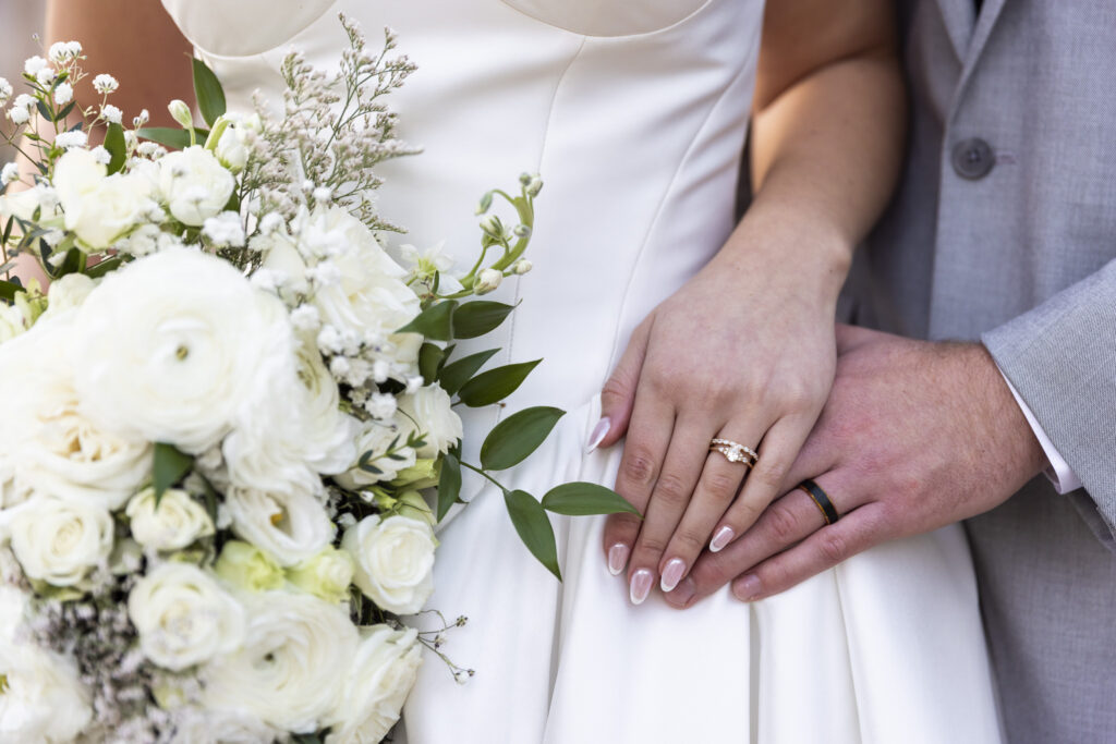 A close-up detail of the bride’s bouquet and the couple’s hands resting together at her waist, her engagement ring and wedding band clearly visible against the smooth fabric of her dress. The groom’s hand overlaps hers, grounding the image in touch and texture