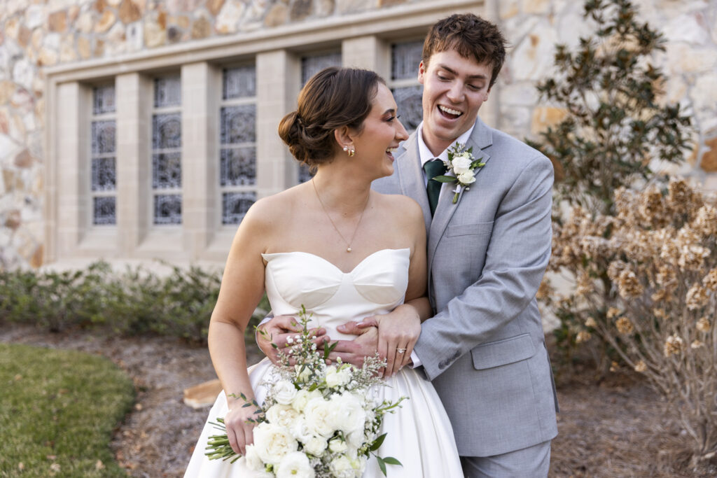 The couple laughs together mid-movement, bodies turned toward each other as the groom wraps his arms around the bride from behind. She holds her bouquet low at her waist, smiling up at him as the church windows and landscaping form a soft backdrop. The relaxed joy captured here reflects the ease and connection felt throughout their Lookout Mountain Presbyterian Wedding.