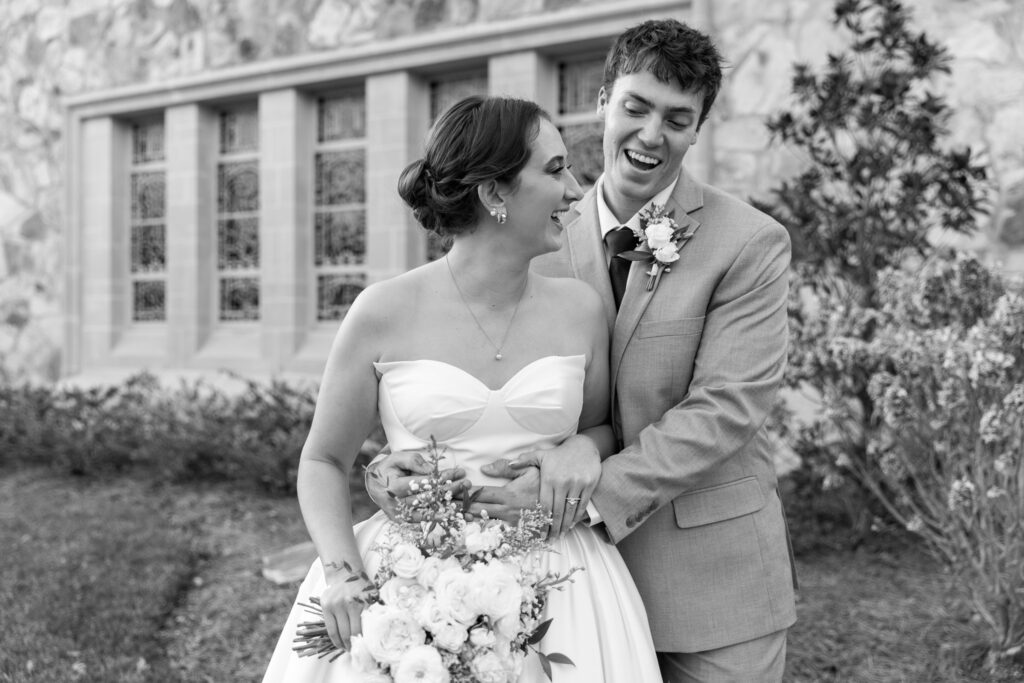 The couple laughs together mid-movement, bodies turned toward each other as the groom wraps his arms around the bride from behind. She holds her bouquet low at her waist, smiling up at him as the church windows and landscaping form a soft backdrop. The relaxed joy captured here reflects the ease and connection felt throughout their Lookout Mountain Presbyterian Wedding.