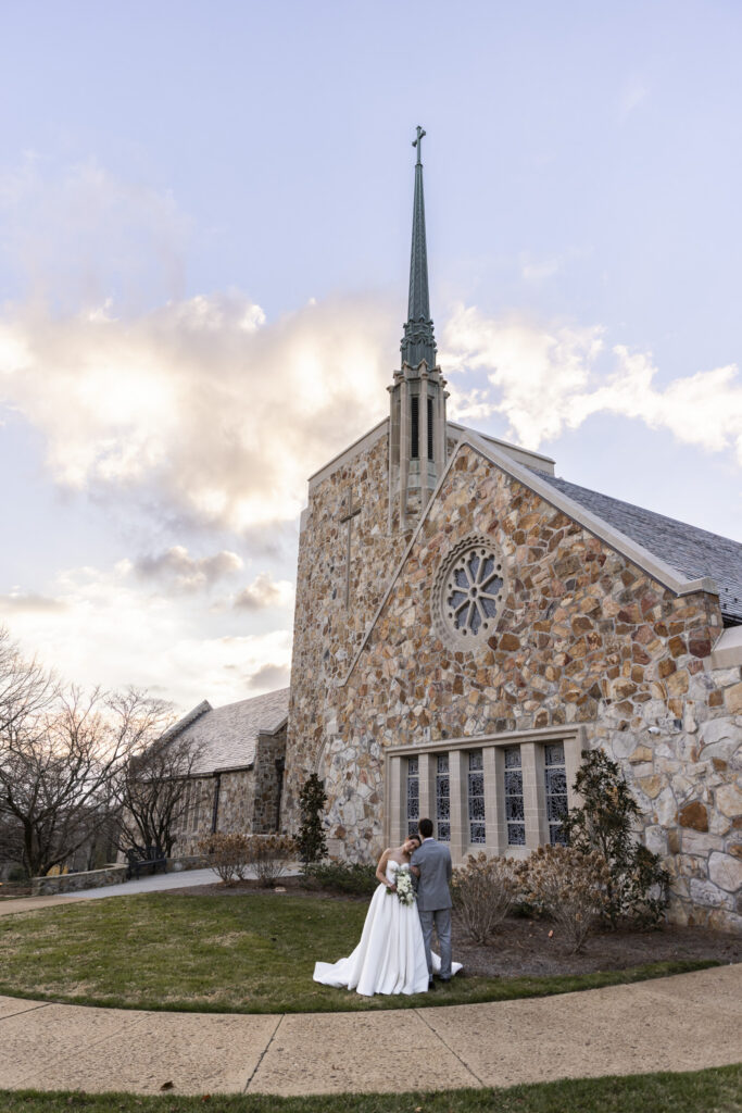An image of the couple standing together outside a stone church, the bride lays her head on the grooms shoulder, while the tall steeple and cross above them. The bride’s full gown and long veil spread softly onto the grass while the groom stands close at her side, bouquet resting against her hip. The scale of the architecture emphasizes the quiet significance of this Lookout Mountain Presbyterian Wedding, grounding the moment in place and tradition.