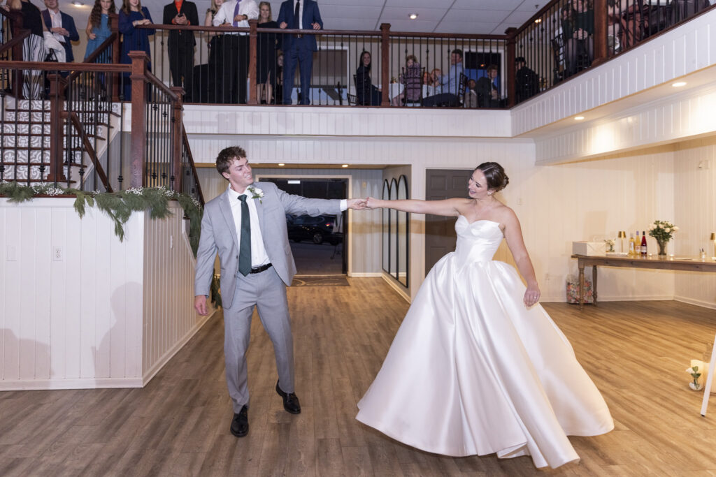 The couple spins playfully across the dance floor, arms extended as they smile at one another mid-movement. Guests line the balcony above, watching and cheering as the bride’s full skirt fans outward. This joyful, sweeping motion captures the celebratory spirit