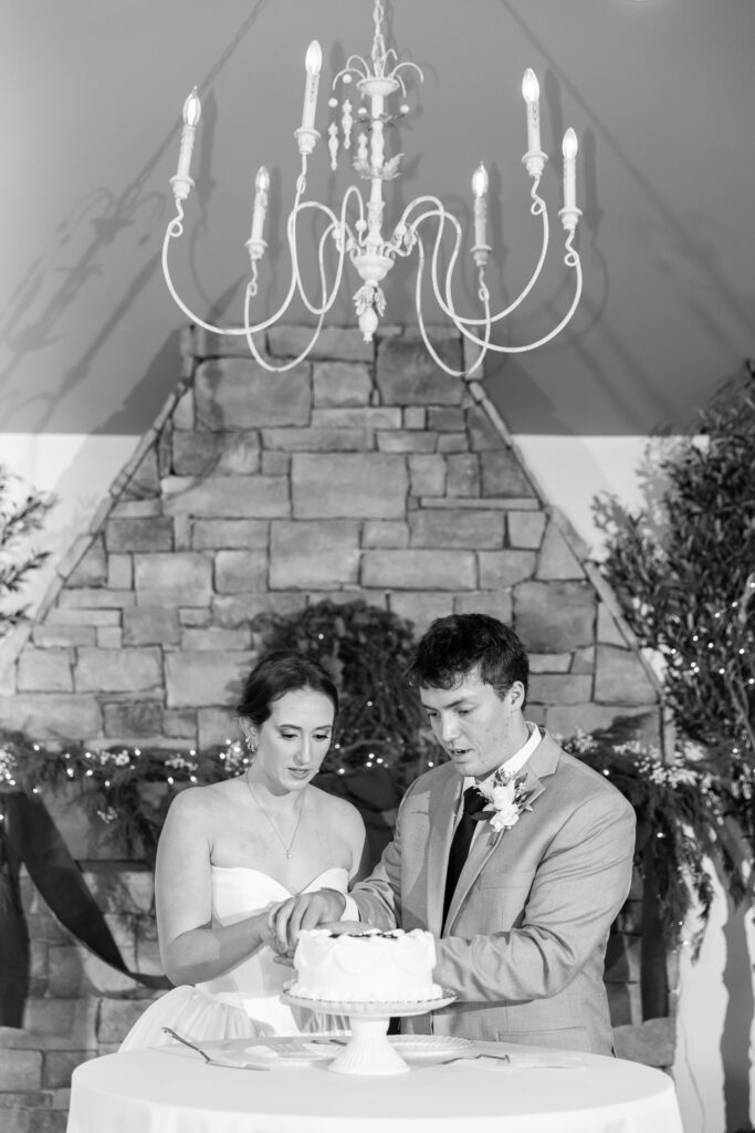 The couple stands side by side cutting their wedding cake together, hands stacked on the knife as they concentrate on the first slice. A stone fireplace and chandelier rise behind them, framing the scene with soft greenery and twinkle lights. This shared moment feels grounded and ceremonial within The Venue Chattanooga wedding reception space.
