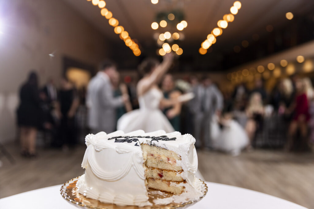 A close-up of a partially sliced wedding cake sits in sharp focus on a table in the foreground, revealing multiple layers and frosting inside. In the blurred background, the couple dances together beneath soft string lights, guests gathered around them. The contrast between the still cake and the motion behind it captures the rhythm of The Venue Chattanooga wedding reception.