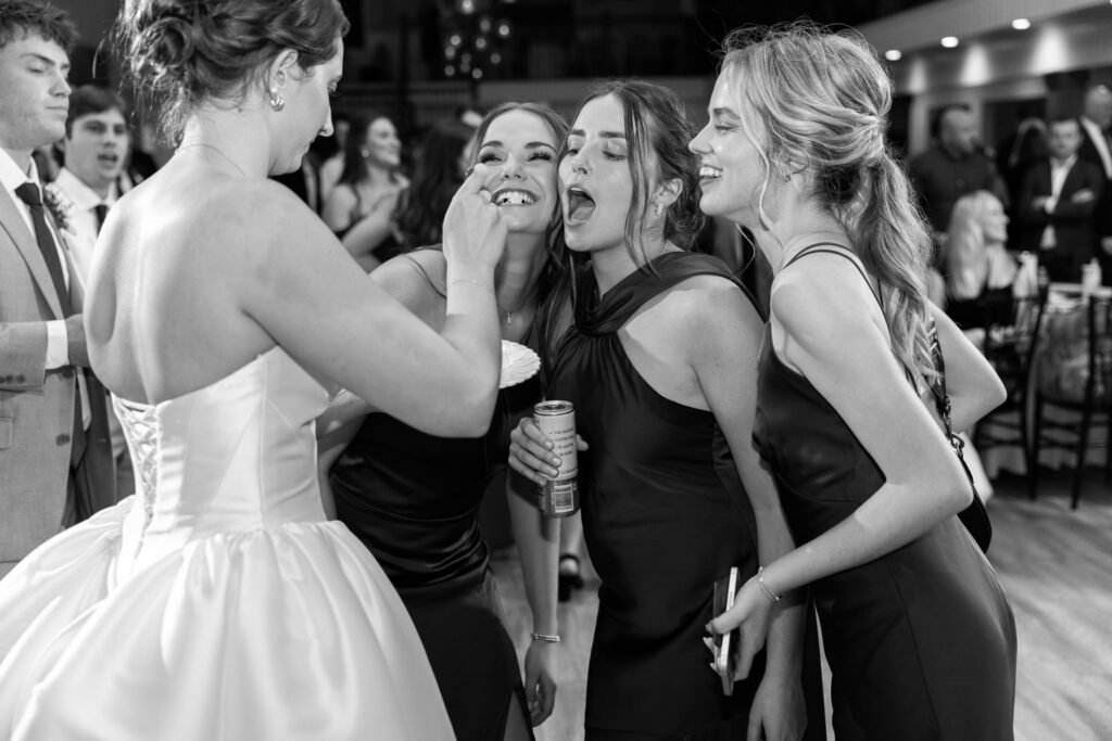 The bride feeds cake to three friends at once, holding a fork forward as the women lean in laughing with mouths open. One bridesmaid holds a drink can while another smiles wide, their dresses dark against the bride’s light gown. This playful, candid interaction unfolds naturally on the dance floor during The Venue Chattanooga wedding.