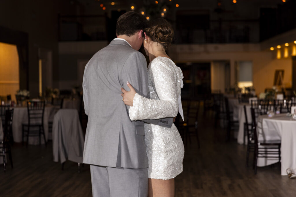 Inside the reception space, the couple stands close together on the dance floor, foreheads touching in a quiet, intimate moment. The groom’s arm wraps around the bride’s back as she rests her hand against his shoulder, her sequined dress catching the light. Empty tables and chairs fade softly into the background, grounding this calm pause during The Venue Chattanooga wedding before the celebration continues.