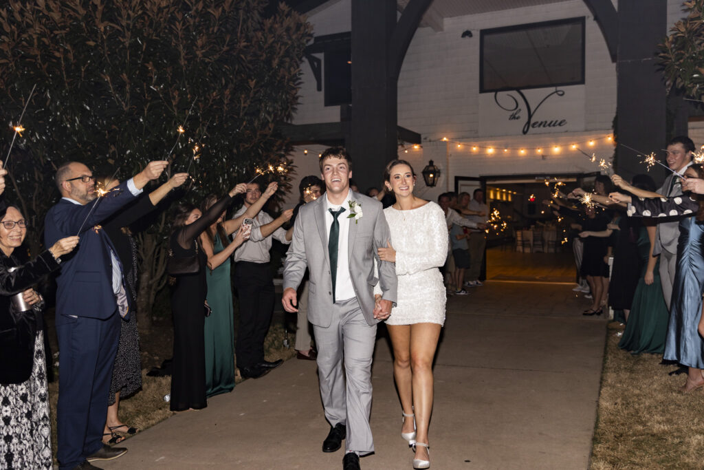 A newly married couple walking hand in hand through a sparkler exit, guests lining both sides of the walkway with arms raised as sparks fly around them. The groom wears a light suit and tie, smiling straight ahead, while the bride wears a long-sleeve, sequined mini dress and heels, her free hand wrapped around his arm. Behind them, the building signage and string lights mark this joyful nighttime moment at The Venue Chattanooga wedding, with laughter and movement filling the frame.