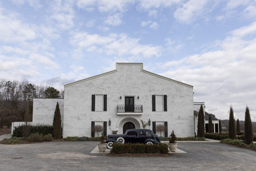 A wide, centered exterior view of a white stucco estate building at The Woodlands at Howe Farms, featuring black shutters, arched brickwork, and a wrought-iron balcony above the main entrance, with a vintage black car parked in front under a soft, cloud-filled sky.