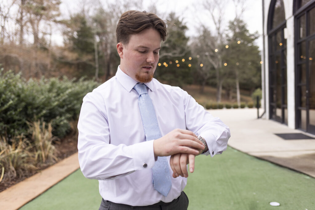 The groom stands outdoors on the paved grounds of The Woodlands at Howe Farms, adjusting his wristwatch while wearing a white dress shirt and light blue tie, with string lights glowing softly behind him and winter trees lining the background.
