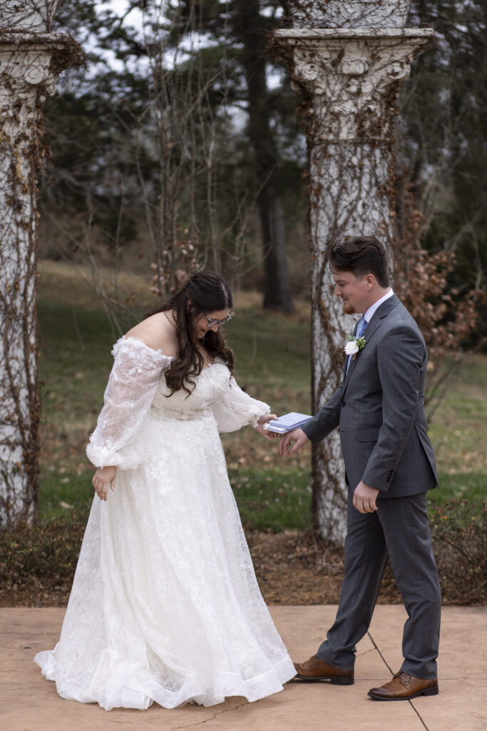 The couple stands facing one another between tall, weathered columns at The Woodlands at Howe Farms, holding hands and looking down with quiet emotion as the bride’s lace gown pools around her feet and fallen leaves scatter across the ground.