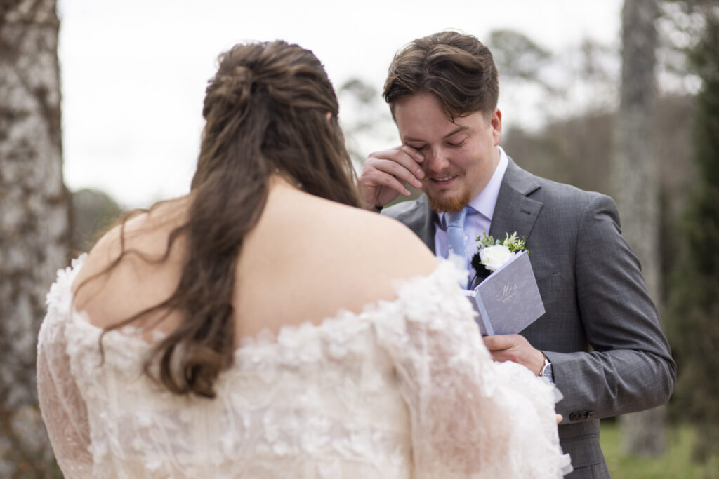 During their first look, the groom wipes a tear while holding his vow book as the bride faces him just out of focus, a tender exchange preserved in these howe farms wedding photos against a soft outdoor backdrop of trees and muted winter tones.