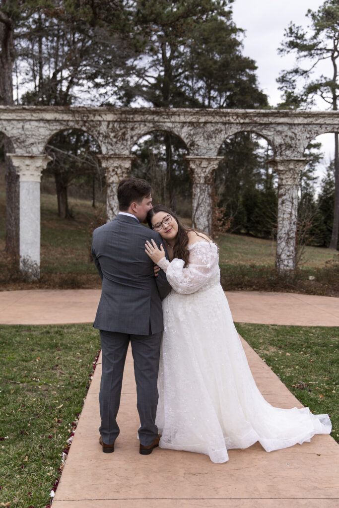 Standing centered on a stone walkway lined with petals, the bride leans her head gently on the groom’s shoulder, eyes closed, as the arches of Howe Farms frame them symmetrically in these howe farms wedding photos.