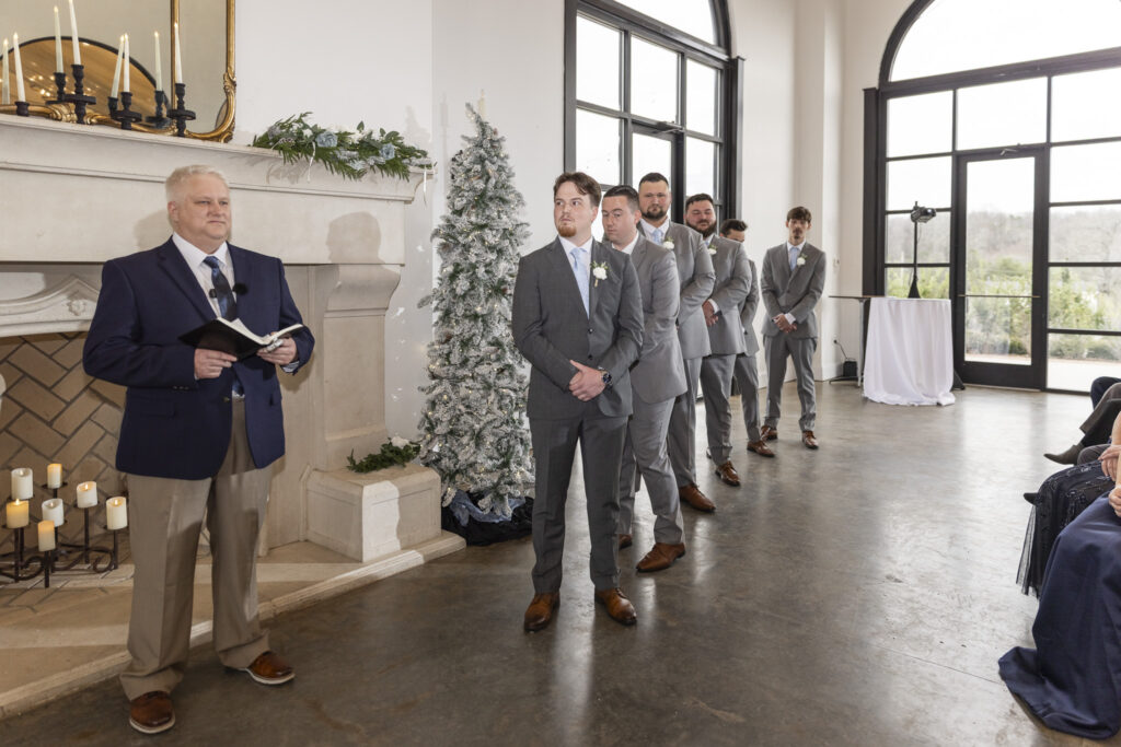 Inside the ceremony space, the groom stands at the altar with his groomsmen lined neatly behind him, an officiant holding a book nearby, while winter greenery and a flocked Christmas tree flank the fireplace in these howe farms wedding photos.